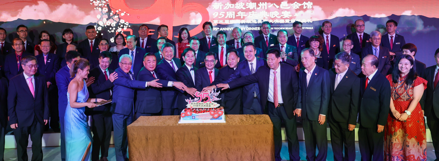 Group of people in suits placing hands on cake at a 95th anniversary dinner celebration.